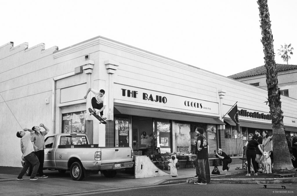 A skateboarder jumping over a car with people watching outside a restaurant. Shot by recently graduated photographer Raymond Alva.